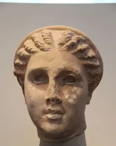 Weathered stone head of Lycosoura Artemis with intricate wavy braids and a headband, mounted on a dark base.