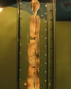 The Shigir Idol stands upright in a glass case, showing an oval head and carved geometric patterns on its weathered wooden surface.