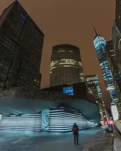 Irish Hunger Memorial with a rocky upper facade and illuminated stepped section beside downtown skyscrapers at night.