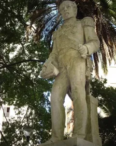 The Count of Porto Alegre monument, a light-colored military figure sculpture with epaulets and a sash, seen from below among trees and palm fronds.