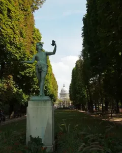 The Greek Actor statue on a tall stone pedestal in a tree-lined park, with a grassy avenue and distant domed building behind it.