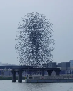 Quantum Cloud sculpture of thin dark metal rods forming a large oval shape beside the water.