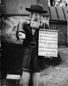 Rosenbom, a wooden bearded figure in a hat holding out one hand and a sign.