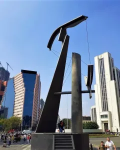 Puerta 1808, a tall angular metallic sculpture, stands in a busy urban plaza with pedestrians, high-rise buildings, and cranes in the background.