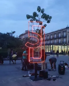 Antimonumento 5J glowing orange in a public plaza at twilight, with people and audio equipment nearby.
