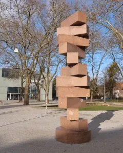 Endlose Treppe by Max Bill, a tall reddish-brown stone sculpture on a paved plaza with trees and a modern building behind it.