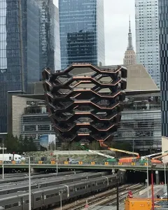 The Vessel sculpture in Manhattan, with honeycomb-like stairways beside glass skyscrapers and train tracks in the foreground.