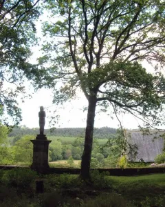 The Vénus de Quinipily stone sculpture stands on a tall pedestal between two trees, with fields and forest in the background.