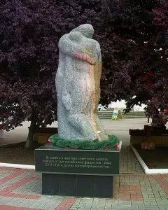 The Shot in the Back monument showing two stone figures on a dark base with Cyrillic text, set among trees and paved paths.