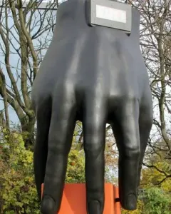 Hand with Clock sculpture on a tall pedestal beside a digital time display, with trees, bicycles, and a nearby building in the background.