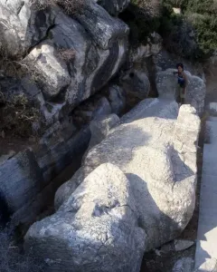 The unfinished Kouros of Apollonas лежing in a quarry, with a person standing beside its massive stone form.