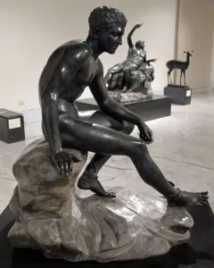 Seated Hermes bronze statue of a nude male figure on a rock, looking downward.