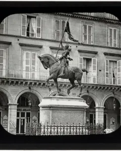 Joan of Arc equestrian statue on a tall pedestal in front of a building with arched shops and shuttered windows.