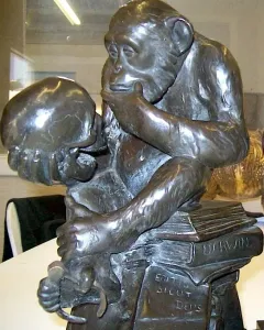 Bronze sculpture of a chimpanzee holding a human skull while seated on books, including one labeled Darwin.