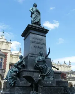 Adam Mickiewicz Monument with a bronze standing figure on a tall pedestal, framed by historic buildings and a blue sky.