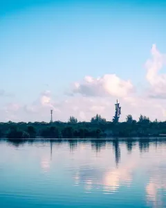 Birth of the New World sculpture reflected in calm water near a tree-lined shore.