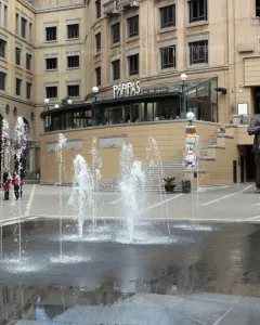 Nelson Mandela statue with raised arms in a busy public square, standing behind a fountain.