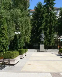 Monument to Hristo Botev: a bronze-colored statue of a bearded man holding a rolled document on a tall stone pedestal among trees.