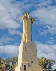 Monument to Columbus, a tall stone sculpture on a rectangular base in an open plaza with trees and a blue sky.