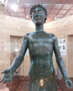 Efebo de Selinunte, a dark bronze nude young man statue with one arm raised in a museum display.