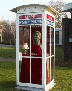 A Stalin bust displayed inside a glass emergency box on a grassy lawn, with a small lamp and fish in front.