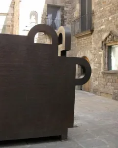 Topos V, a dark geometric sculpture with cutouts, stands in front of historic stone buildings in Barcelona.