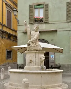 Dolphin Fountain with Triton, a stone sculpture of a bearded man holding a fish in a cobblestone square.