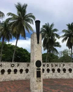 The Torch of Friendship monument’s stone pillar topped with a dark urn and small flame, with palm trees and a low wall behind it.