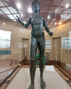 Efebo de Selinunte, a dark bronze nude young man statue with one arm raised in a museum display.