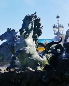 Bronze fountain at the Monument aux Girondins with rearing horses and spraying water, with a fairground attraction and ornate lamppost in the background.