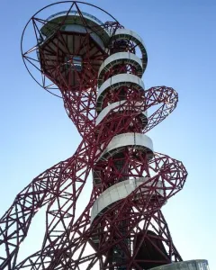 Orbit Tower’s red metal framework with a white spiral walkway rising to the top against a blue sky.