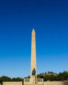 National Women\'s Monument obelisk with a bronze woman and child sculpture at the base, framed by trees and shrubs.