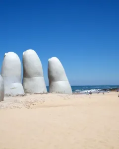 The Hand sculpture rising from the sand on a beach with the ocean and blue sky in the background.