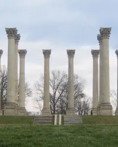 Capitol Columns standing in a wide arc on a grassy field with steps in the foreground and cloudy skies above.
