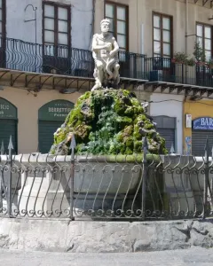 The Fountain of the Genius of Fieravecchia in a city square, with a bearded marble statue on a moss-covered rocky mound behind a protective metal fence.