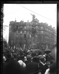 The Monument to Mickiewicz stands in a crowded Paris square during its inauguration, with a statue atop a decorated column and spectators in hats and umbrellas below.