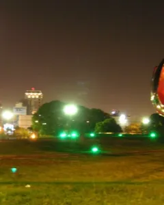 Floralis Genérica partially closed at night, lit by green ground lights with city lights and a bright moon behind it.