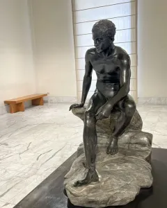Seated Hermes bronze statue of a nude male figure on a rock, looking downward.