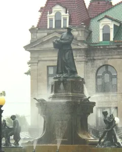 La Fermière bronze statue atop a fountain with water jets, with smaller figures at the base and a historic building behind it.