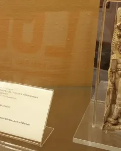 Ivory Lakshmi statuette with elaborate jewelry, headdress, and carved figures at the sides, displayed upright in a clear case.