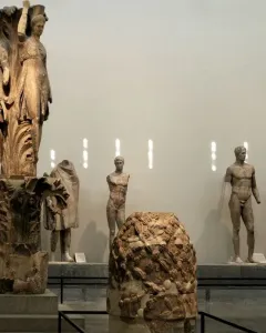Dancers of Delphi sculpture with three draped female figures on a carved column base in a museum hall.