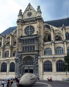 Écoute, a pale stone head sculpture with a hand cupped to its ear, stands in a plaza with a church behind it.
