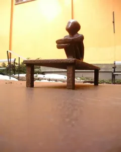 Järnpojke, a small dark seated sculpture on a low bench beside a building wall with patches of snow and greenery.