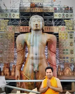 Gommateshwara Bahubali statue with ritual streaks, framed by a decorated scaffolding structure and a smiling man in orange robes in the foreground.