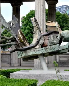 Humpty Dumpty Machine bronze sculpture with a green and brown patina, shown outdoors on a plinth beside classical columns.