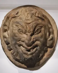 White circular relief sculpture of the Head of a Faun with a grinning face, pointed ears, and curly beard and hair.