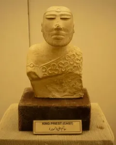 Bust of the Priest King sculpture with a decorative headband and patterned cloak on a dark base
