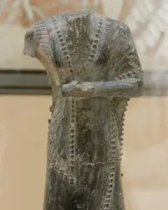 Iddi-Ilum statuette of a headless standing figure with folded arms and a decorated robe on a square base.