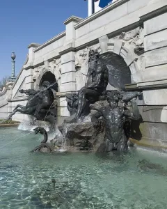 Court of Neptune Fountain with dark metal sea deities and horses rising from a water pool beside an ornate stone building.