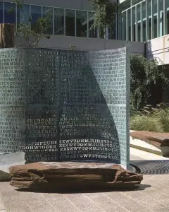 Kryptos bronze sculpture covered with carved capital letters, standing outdoors near stone benches and trees.